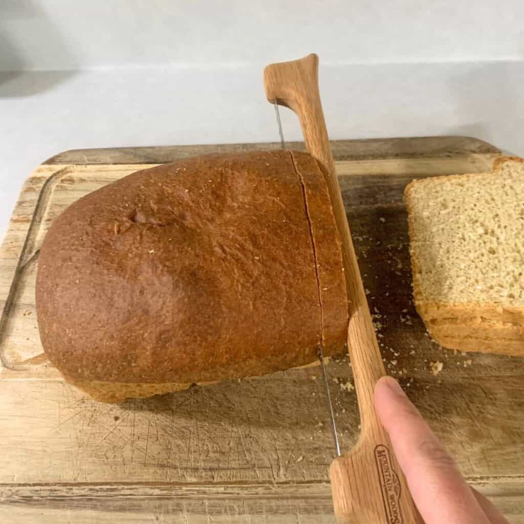 Woman cutting a fresh loaf of sandwich bread on a wooden cutting board using a wood fiddle bow bread knife.