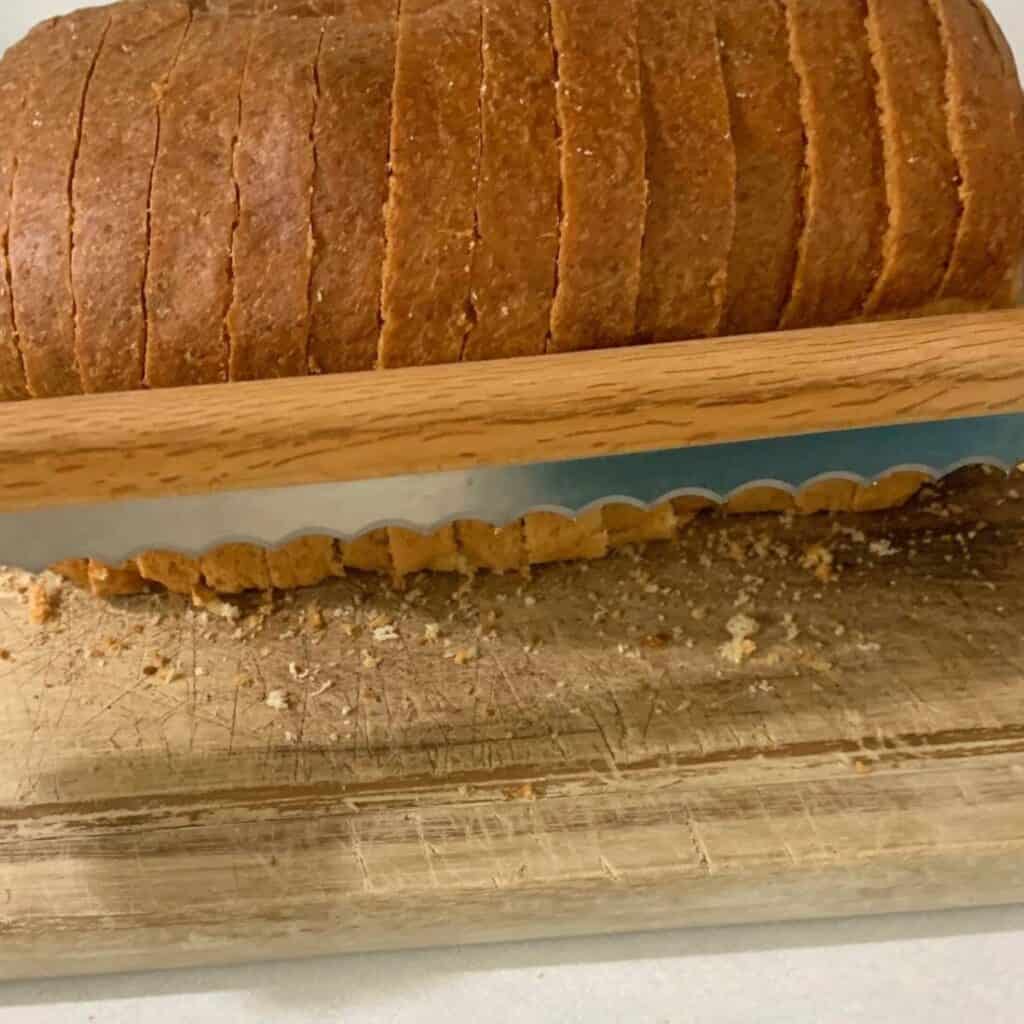 A freshly made loaf of easy sandwich bread on a wooden cutting board. The loaf has been sliced thin with a fiddle bow bread knife. A woman is holding up the fiddle bow bread knife to show the serrated blade.