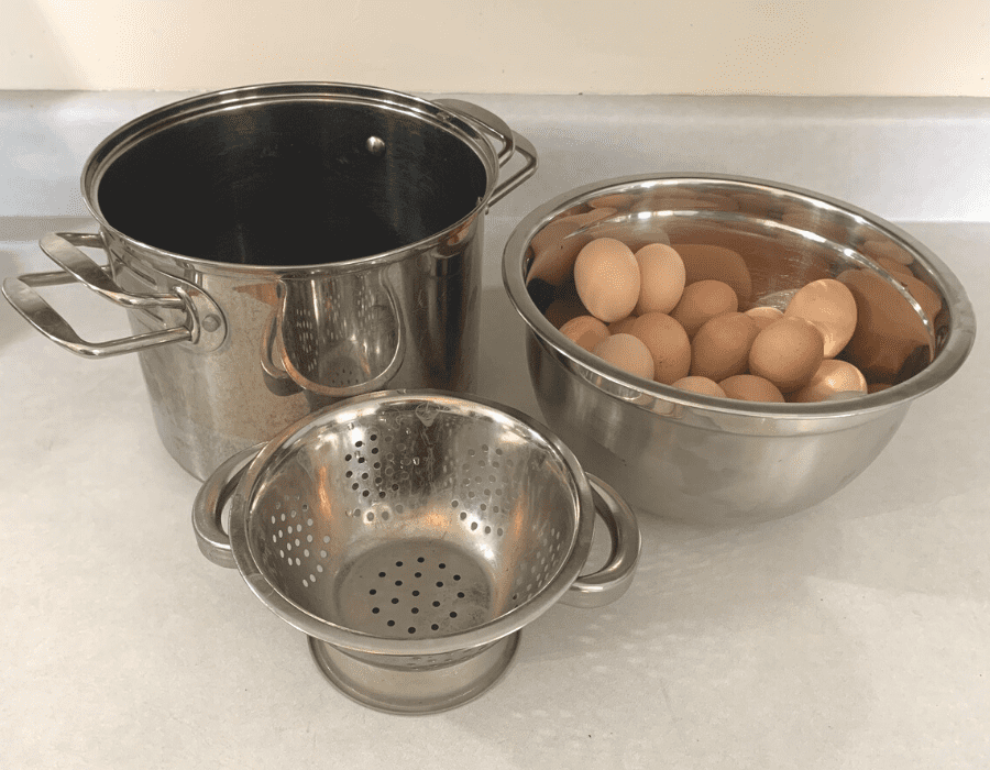 A large stainless steel pot, a small stainless steel colander, and a stainless steel bowl of eggs sitting on a kitchen counter.