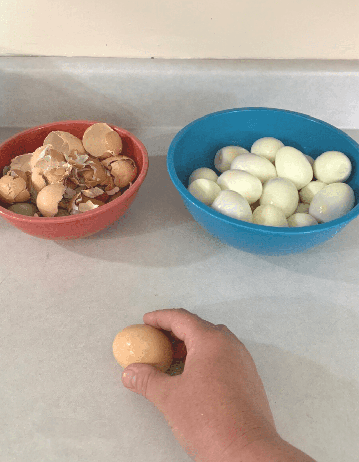 A red bowl of egg shell peelings, a blue bowl of peeled hard boiled eggs, and a woman holding an egg with a brown shell.