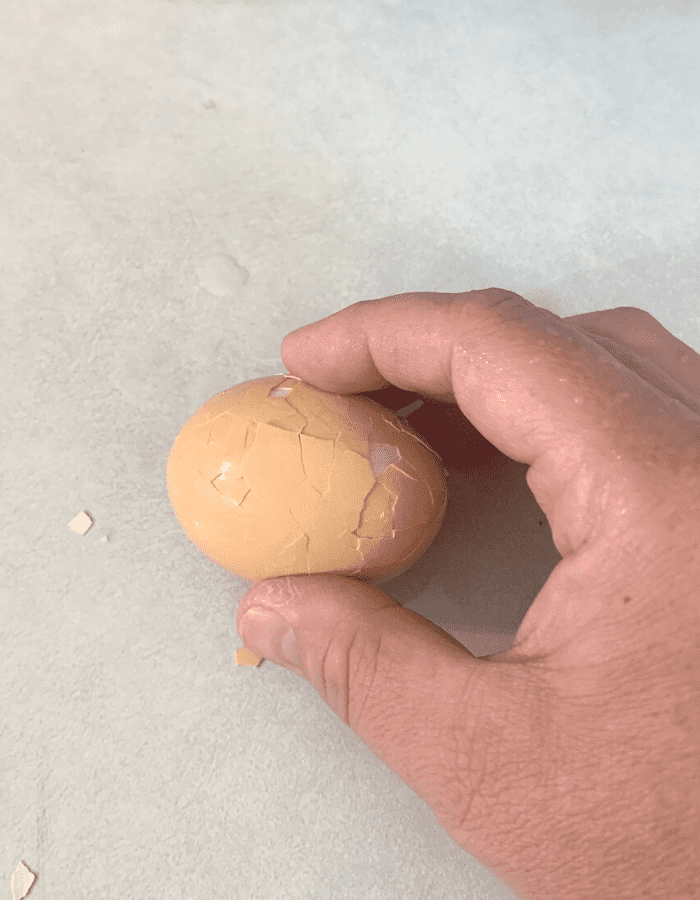 A woman holding a hard boiled egg with a brown shell and crushing the shell on a kitchen counter. She is showing how to peel a hard boiled egg.