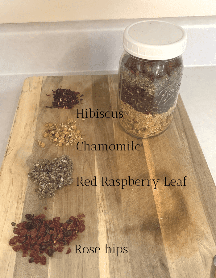 A quart mason jar filled with homemade herbal tea sitting on a wooden cutting board. Scoops of red hibiscus flowers, yellow chamomile flowers, green leaves of red raspberry, and red rose hips are also on the wood cutting board.