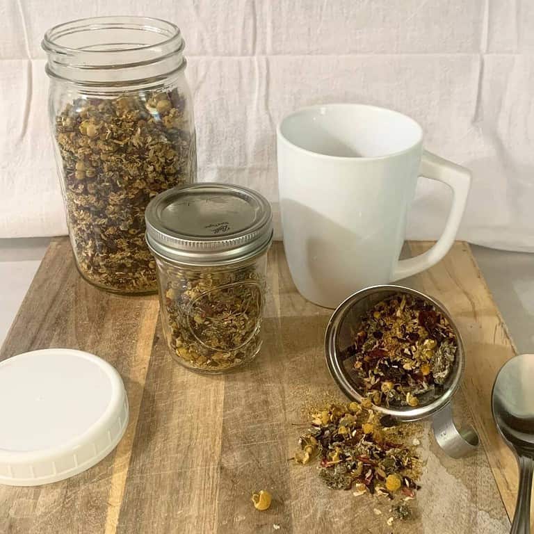 A quart mason jar and half pint mason jar with homemade herbal tea blend sitting on a wooden cutting board next to a white tea cup, white plastic lid, and silver tablespoon. A mesh tea infuser filled with loose leaf tea is spilling out onto the cutting board.