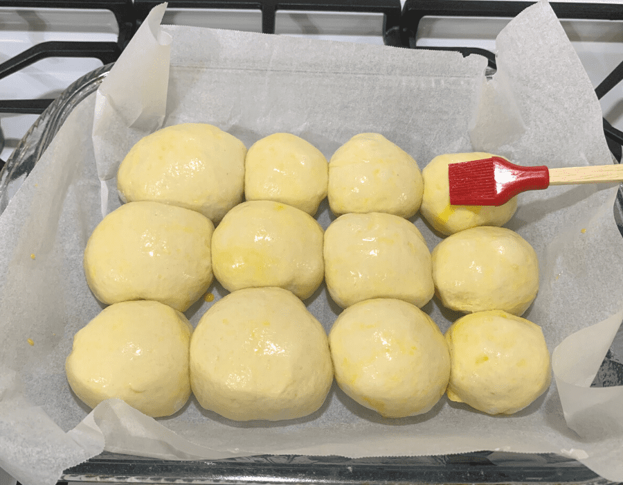 Dinner rolls placed in a baking pan and being brushed with an egg wash.