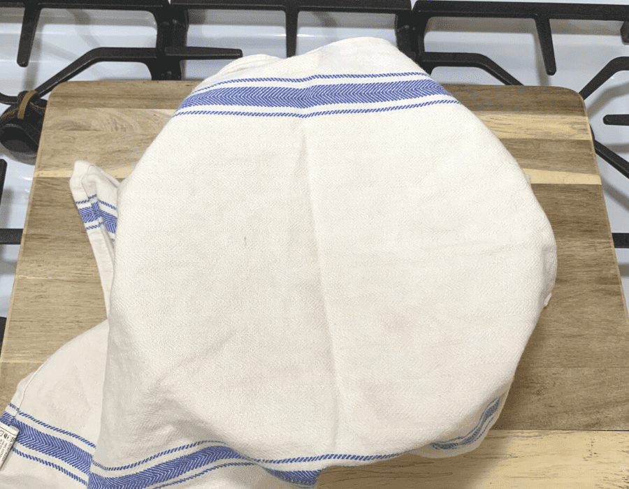 Mixing bowl with bread dough inside with a white tea towel on top.