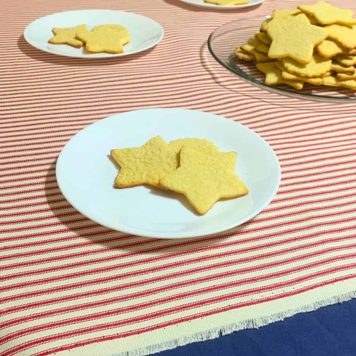 Star-shaped cream cheese sugar cookies on white plates sitting on a table draped with a navy blue tablecloth overlaid with a red and white striped tablecloth.