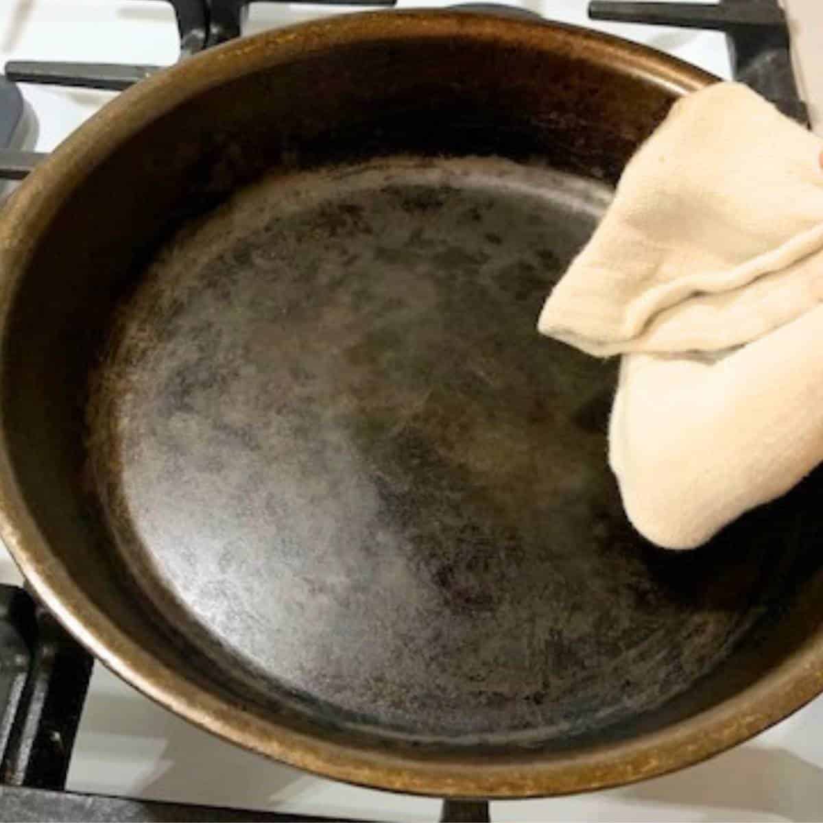 A cast iron skillet on a gas stove with a woman wiping it clean with a white cloth.