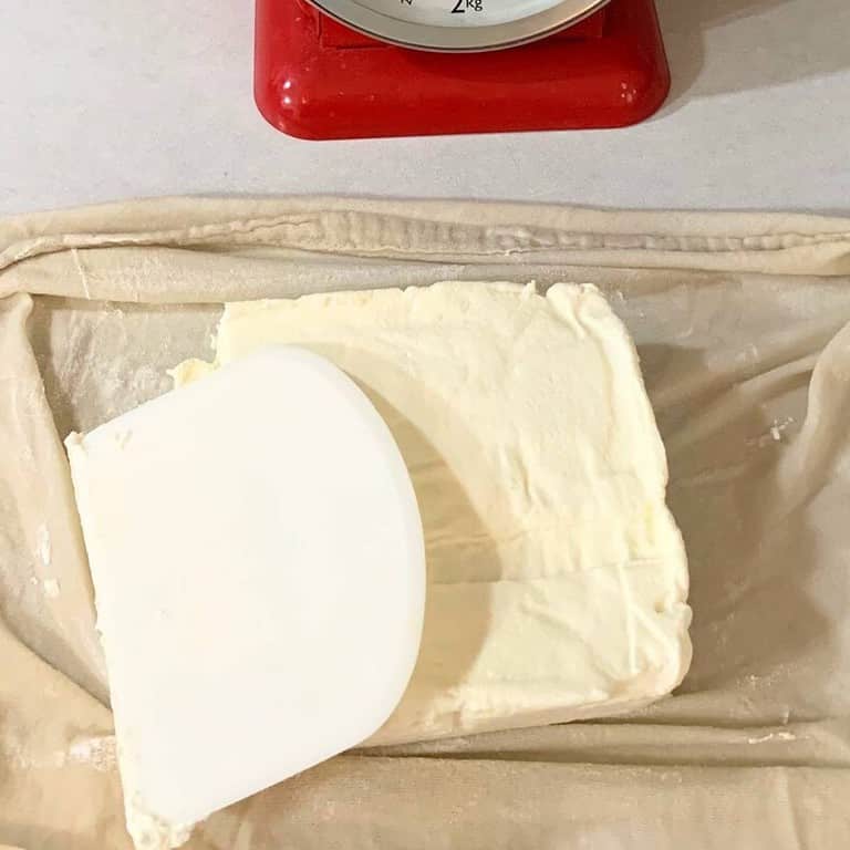 A block of homemade cream cheese on white cheesecloth on a kitchen counter next to a white bench scraper.