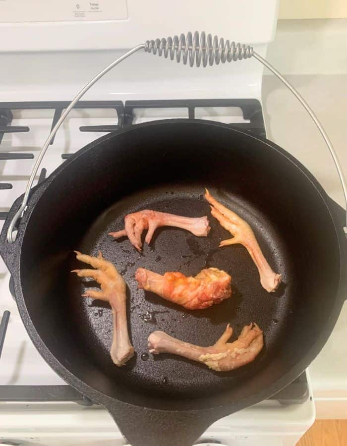A cast iron Dutch oven on top of a stove with chicken feet and chicken neck in the bottom.