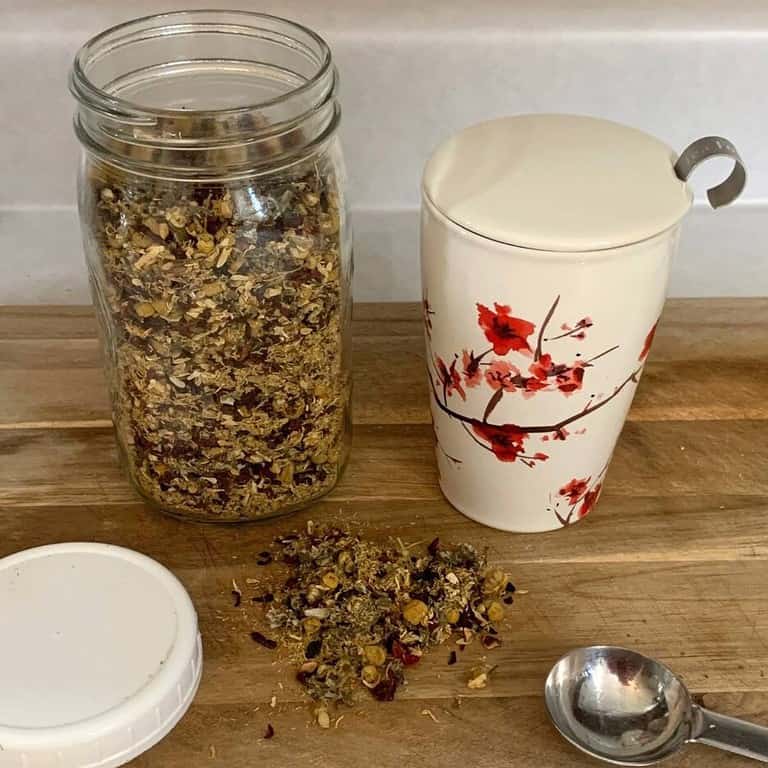 A blend of loose leaf herbal tea in a mason jar and some loose tea on a wooden cutting board. These are next to a tea cup with a lid and a teaspoon.