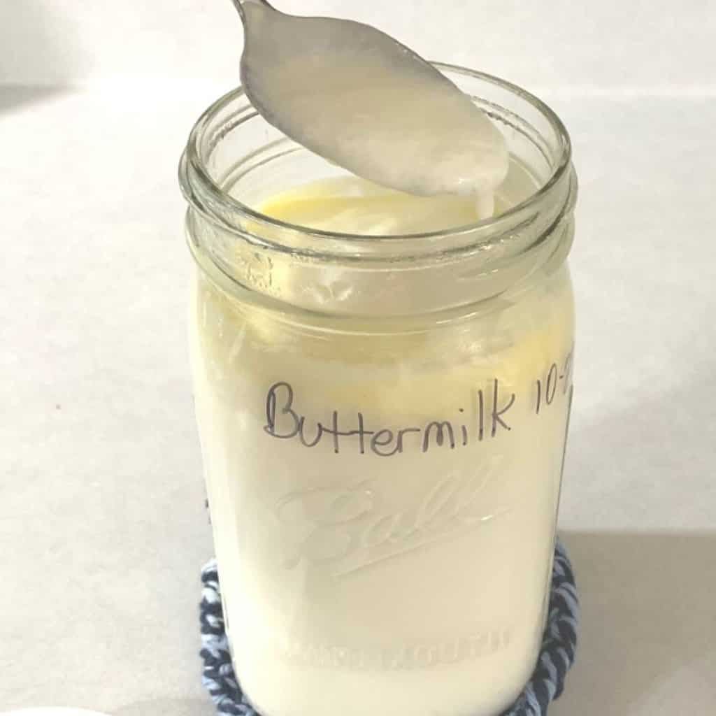 A quart size mason jar of homemade buttermilk sitting on a blue knit coaster on a kitchen counter. A silver spoon has been dipped in and thick buttermilk is dripping off the spoon into the mason jar.