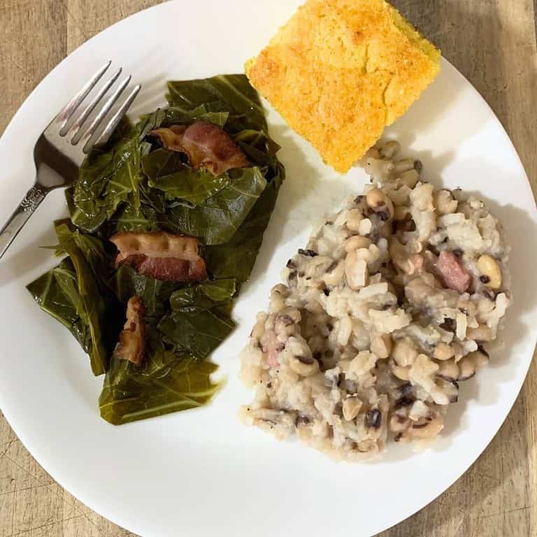 Southern black eyed peas with ham pieces and collard greens with bacon topping next to yellow cornbread on a white plate with a silver fork.