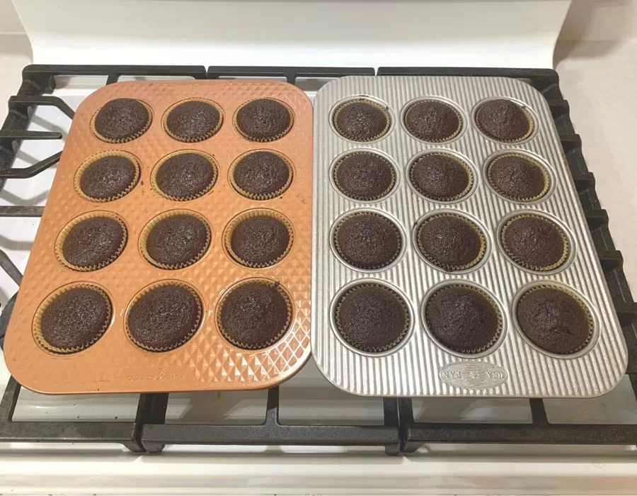 Two dozen chocolate cupcakes freshly baked in muffin pans sitting on top of a stove.