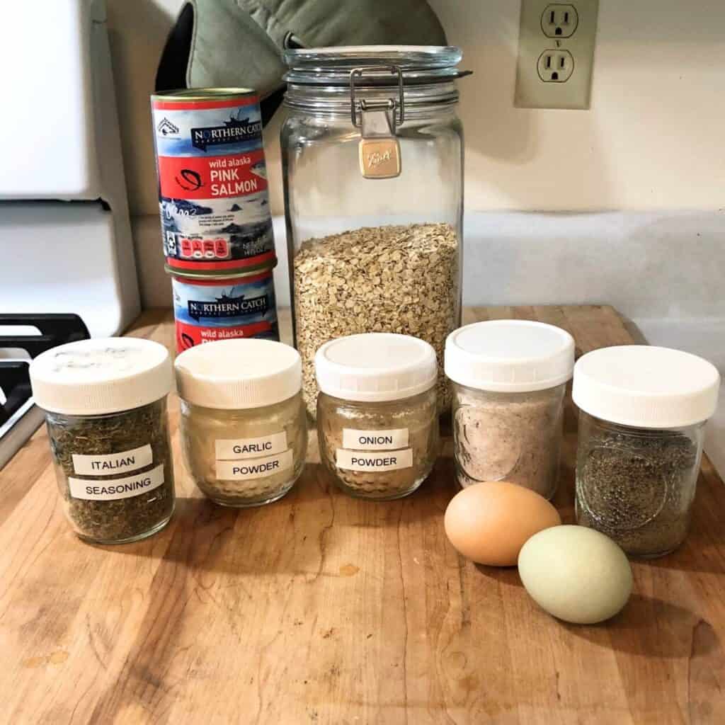 Ingredients on a counter including two cans of wild alaska pink salmon, oatmeal, Italian seasoning, garlic powder, onion powder, salt, pepper, and two eggs.