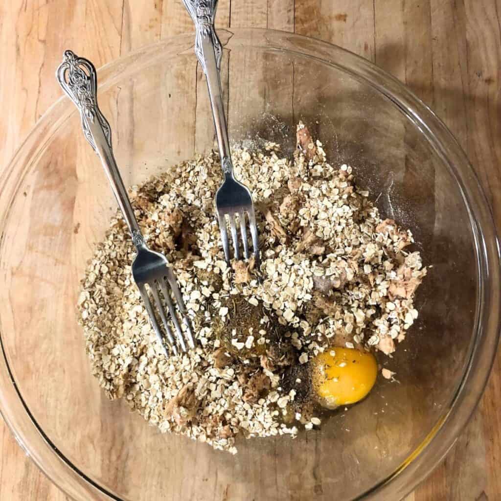 Glass bowl on a wooden counter that contains salmon, oatmeal, eggs, spices, and herbs. Two forks are inside the bowl.