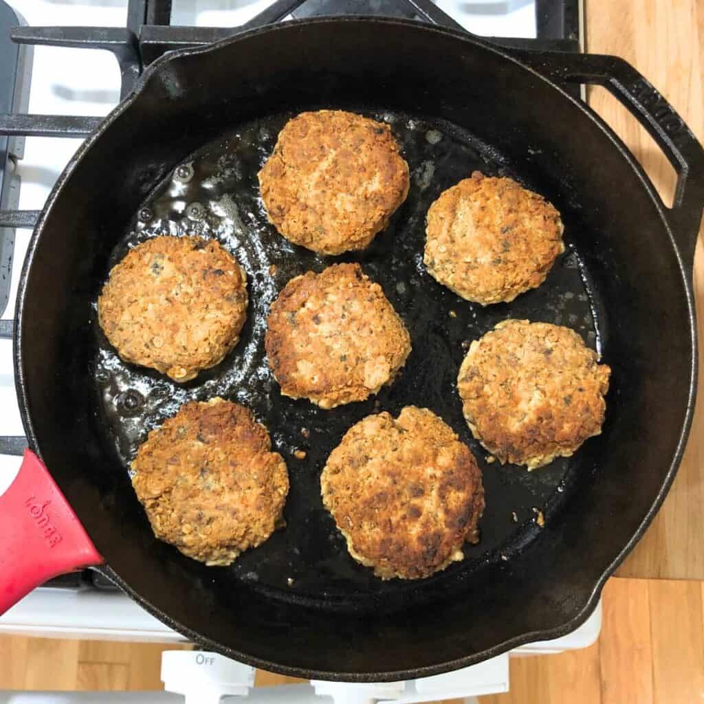 Salmon patties frying in coconut oil in a cast iron skillet on the stove.