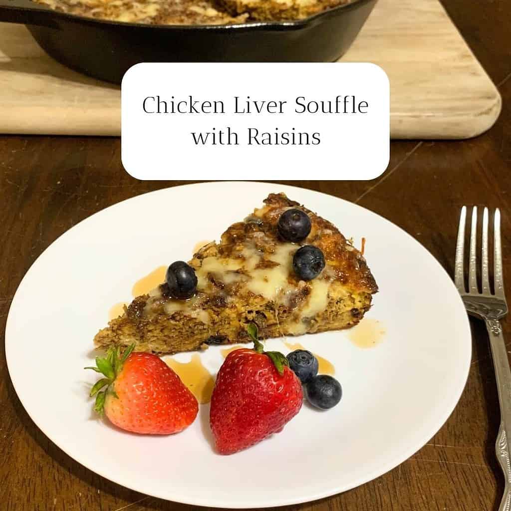 A slice of chicken liver souffle with blueberries and strawberries on a white plate on a dark kitchen table. The title is "Chicken Liver Souffle with Raisins."