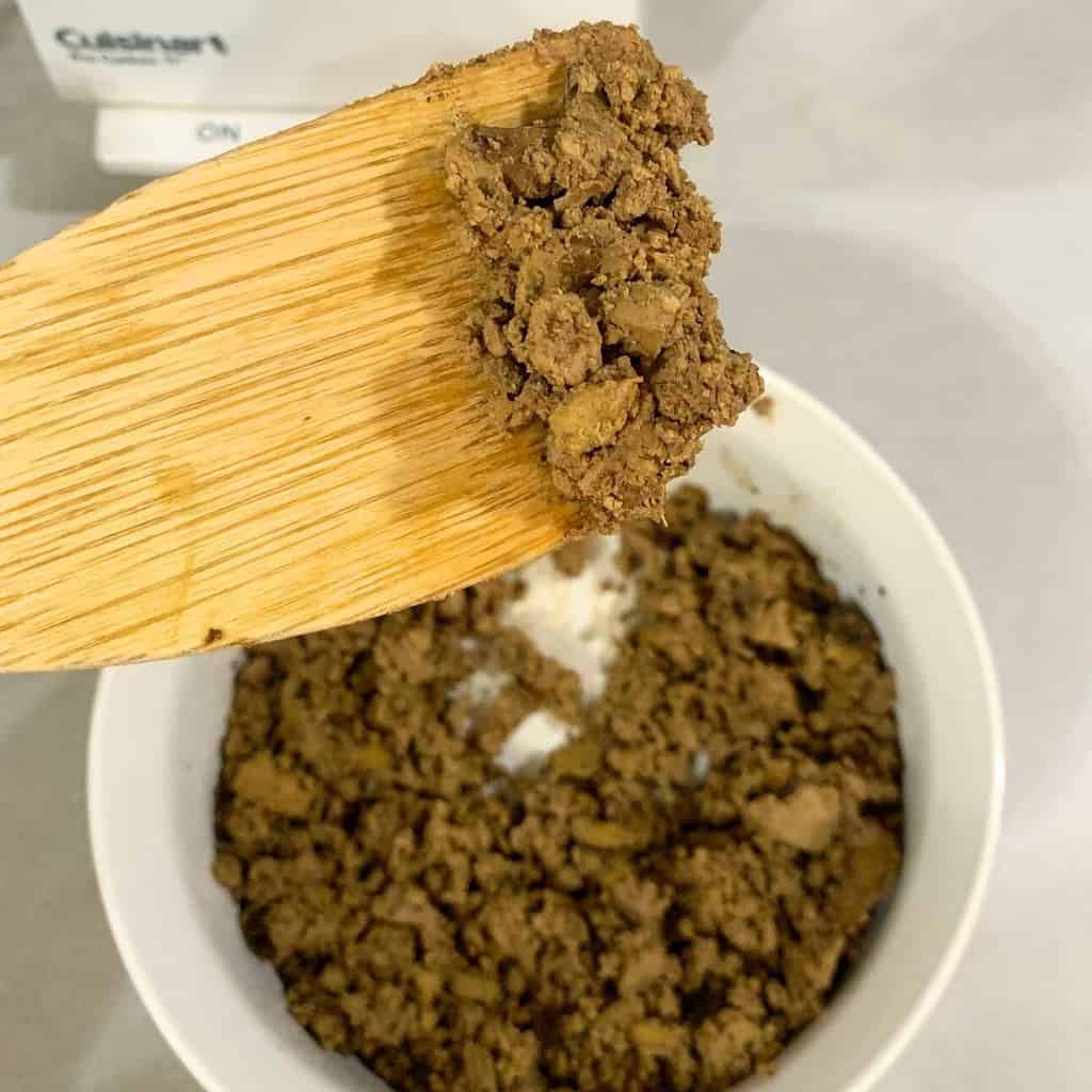 Woman holding a wooden spoon of cooked chicken liver crumbles over a white bowl on the counter.