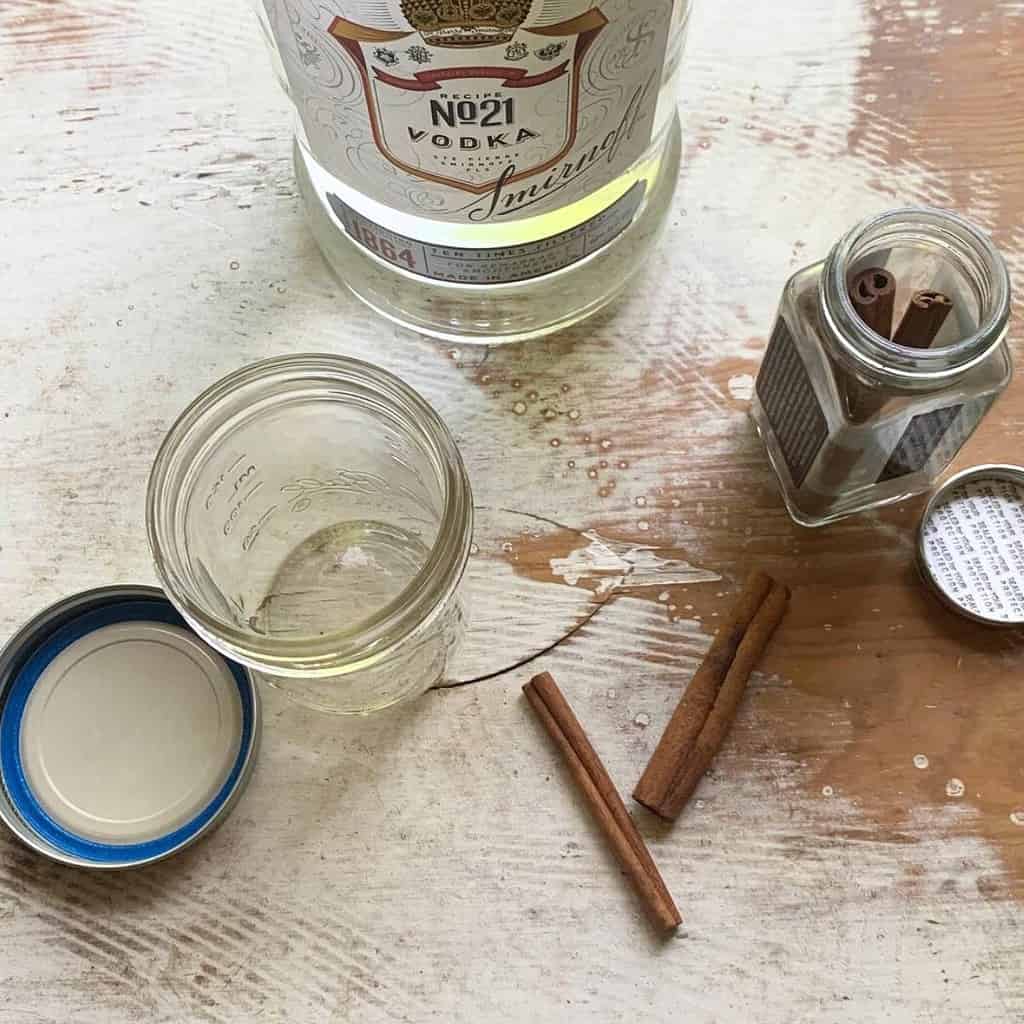 Ingredients to make cinnamon extract on a chipped, white dresser. A glass jar with lid, cinnamon sticks, and a bottle of vodka are sitting on top.