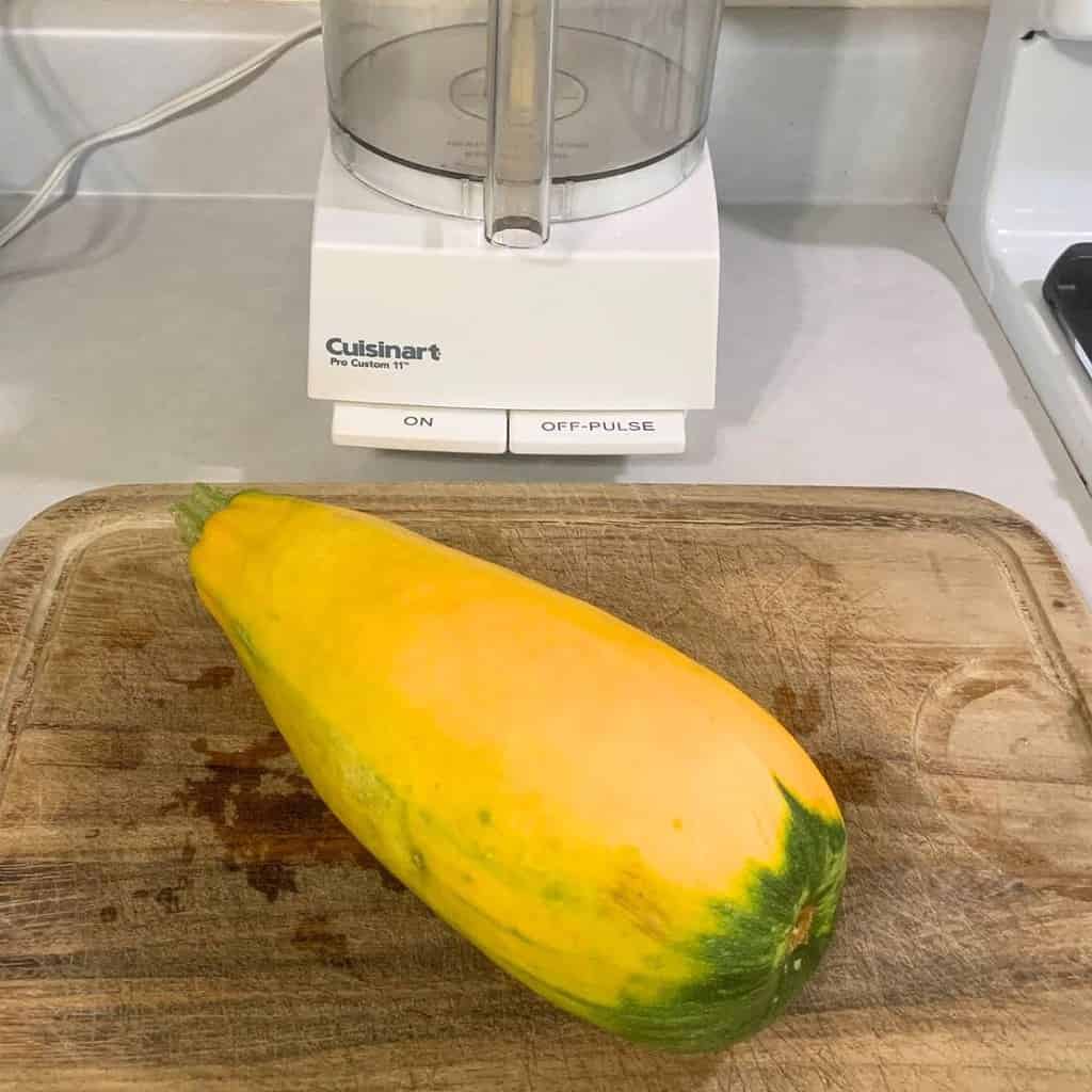 A large golden zucchini on a wooden cutting board in front of a Cuisinart food processor.