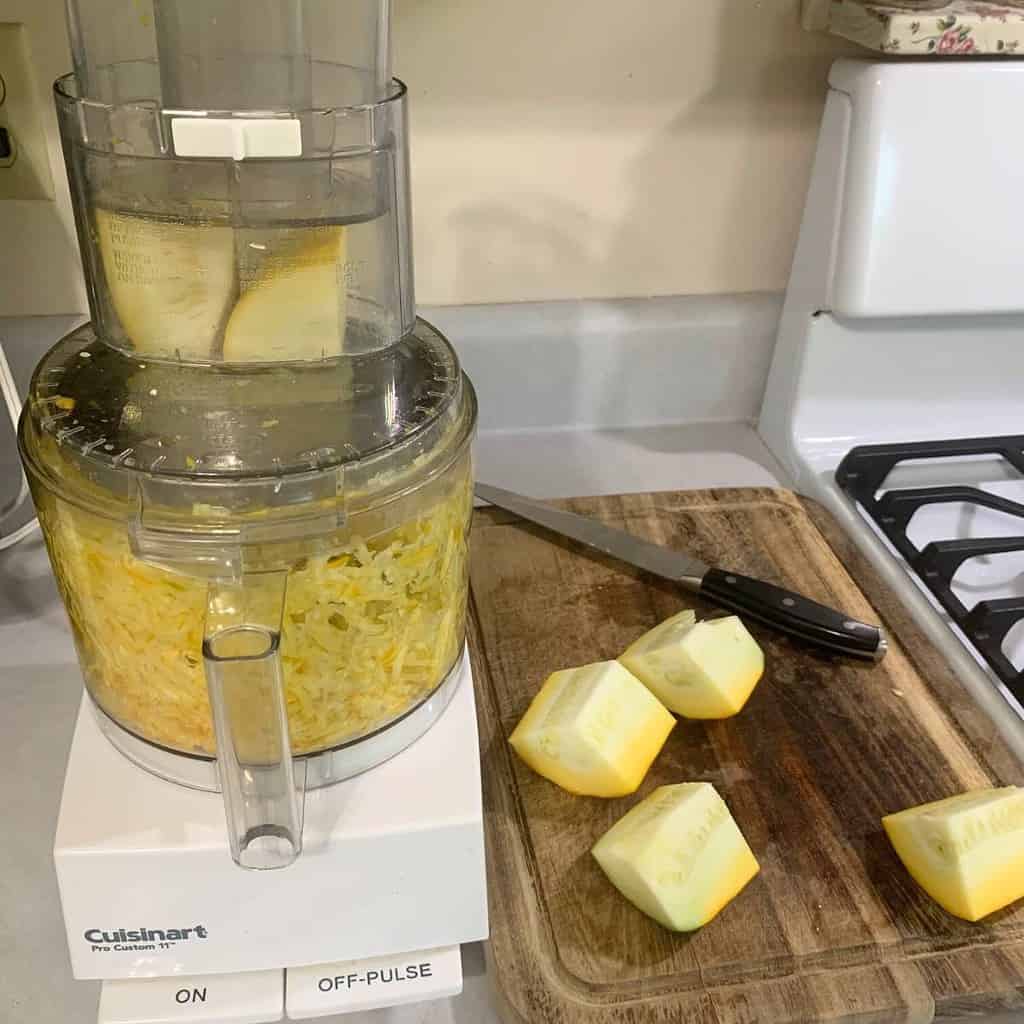 Chopped golden zucchini on a wooden cutting board while zucchini is being grated in Cuisinart food processor.