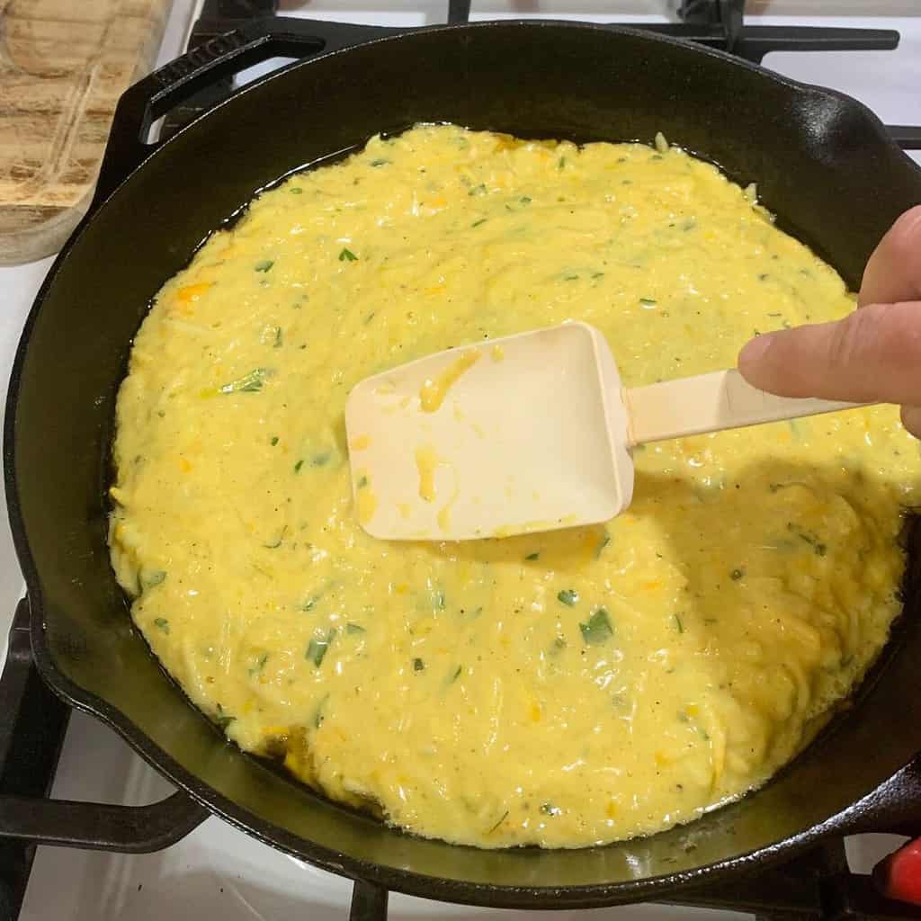 Woman spreading cheesy zucchini casserole in preheated cast iron skillet with a white spatula.