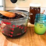 Glass mason jars of raw honey and lemon juice on a wooden cutting board. Next to them is a large glass measuring cup with a fine mesh sieve inserted and soaked herbs are being strained from the liquid.