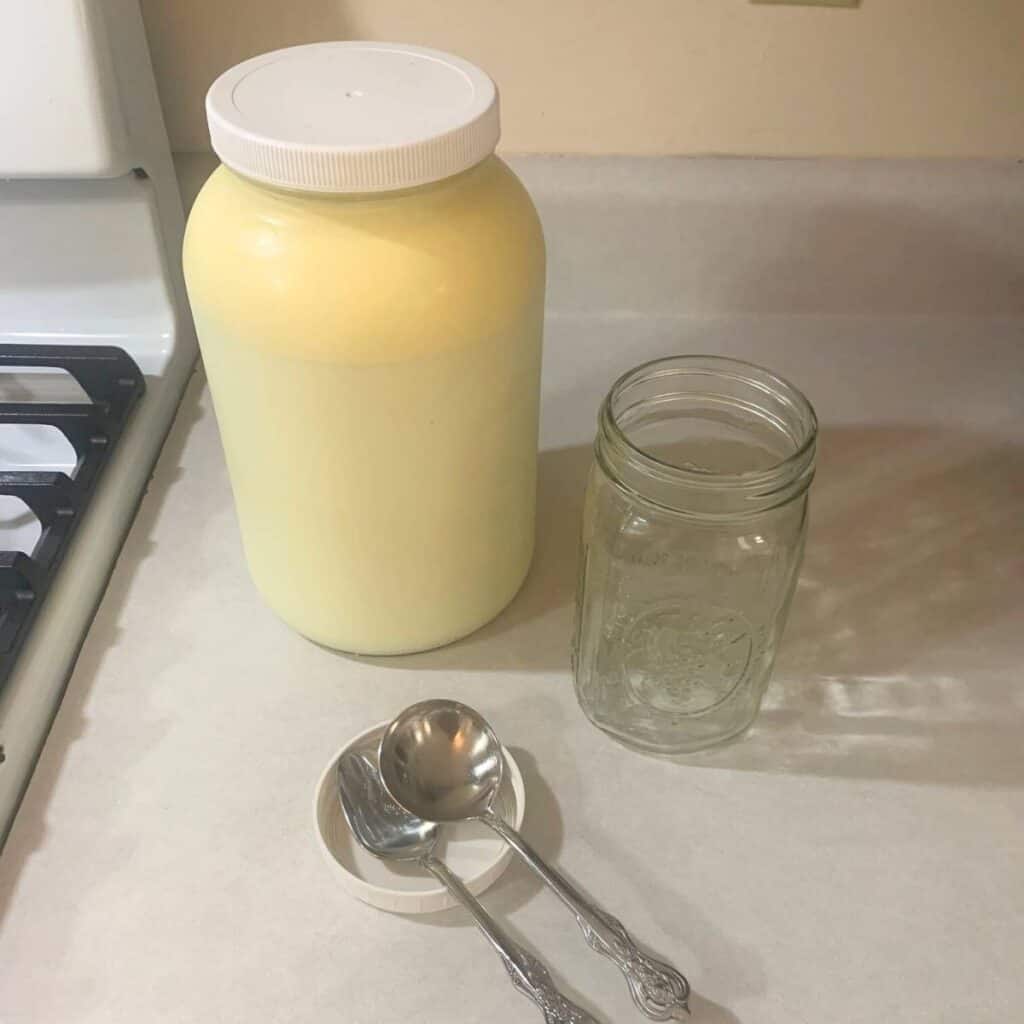 A gallon glass mason jar of raw milk with a separate empty glass quart jar, silver ladle, and silver teaspoon on a kitchen counter.