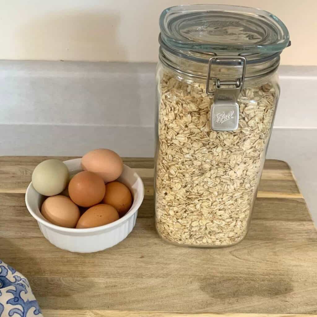 A white bowl with brown and green eggs inside sitting next to a glass mason jar of rolled oats. Both are on a wooden cutting board on a counter.