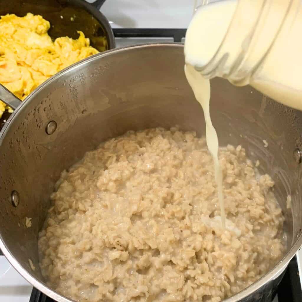 Woman pouring milk from a glass mason jar into a stainless steel pot of cooked oatmeal on the stove. A cast iron skillet of eggs is in the background on the stove.