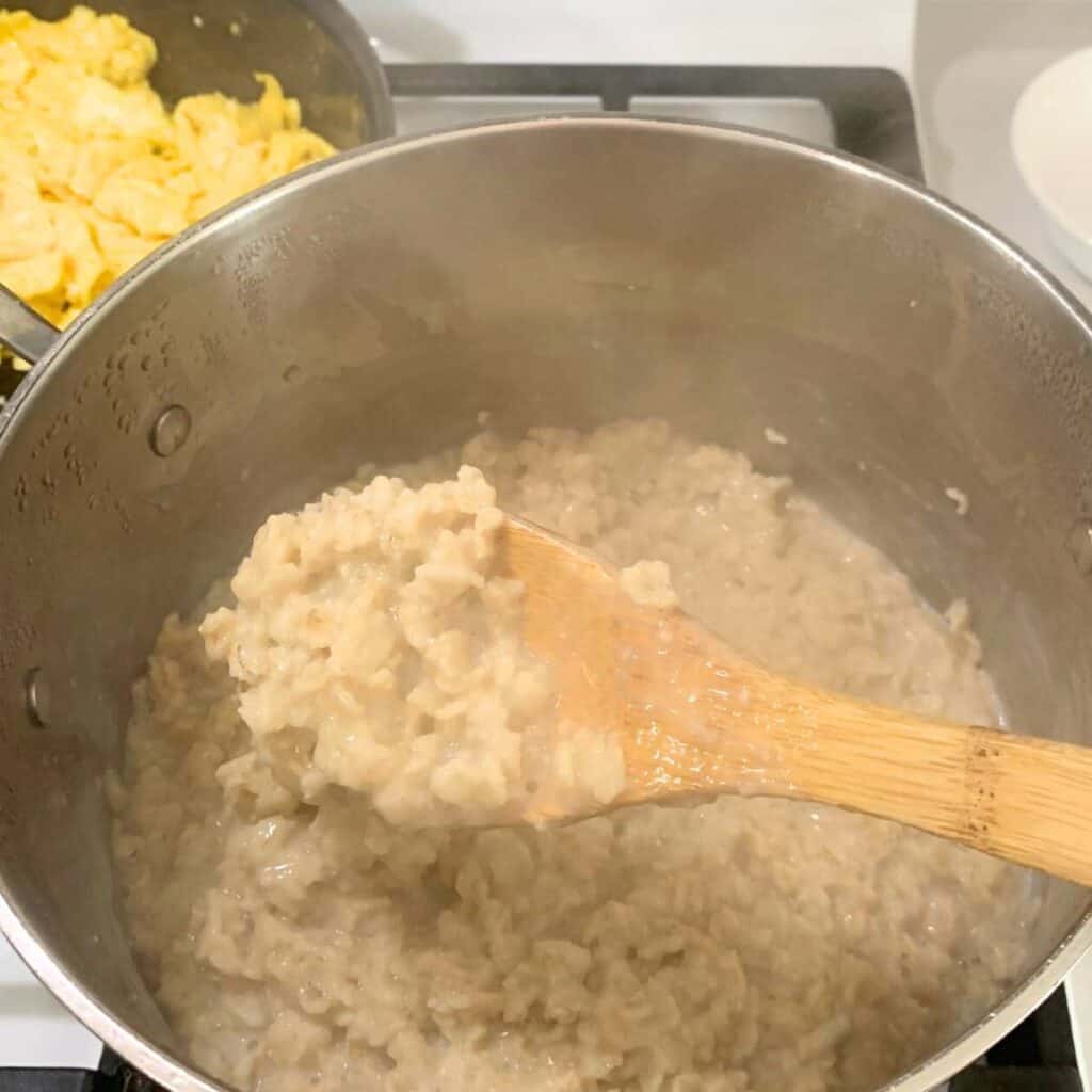 Woman stirring a pot of cooked old-fashioned oats using a wooden spoon. A cast iron skillet of scrambled eggs is behind it on the stove.