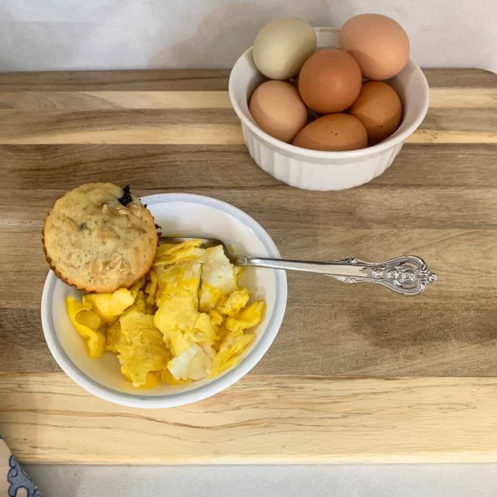 A white bowl of cooked eggs with a blueberry muffin on a wooden cutting board next to a white bowl of green and brown eggs.