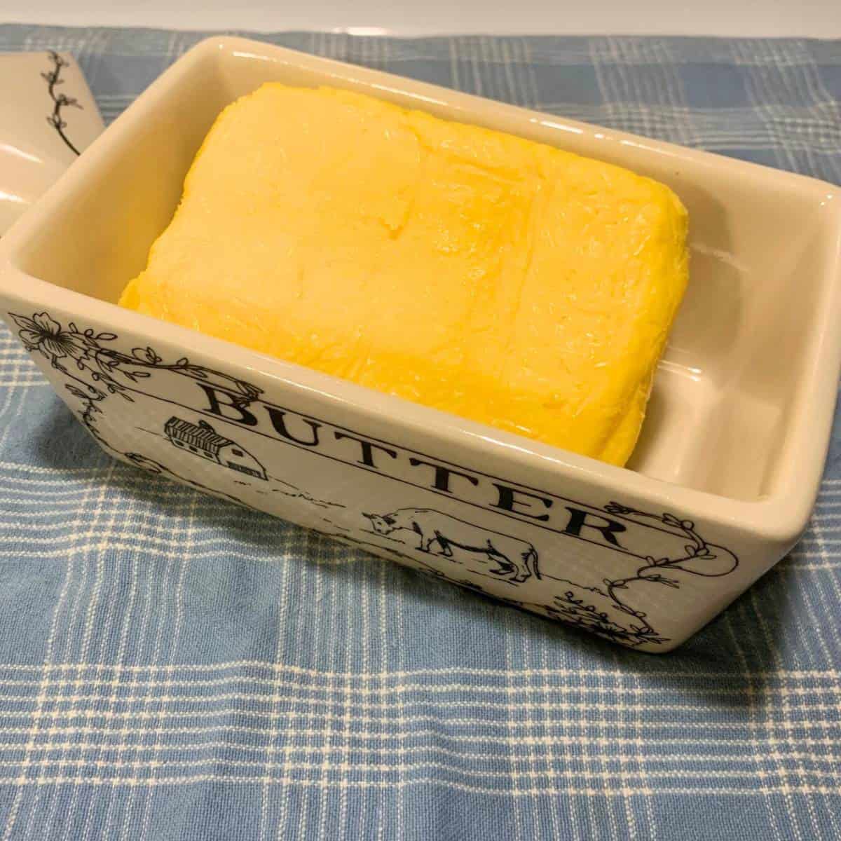 A block of orange-yellow homemade butter in a white ceramic butter dish with black letters that say "BUTTER" with a picture of a cow and barn on the side.