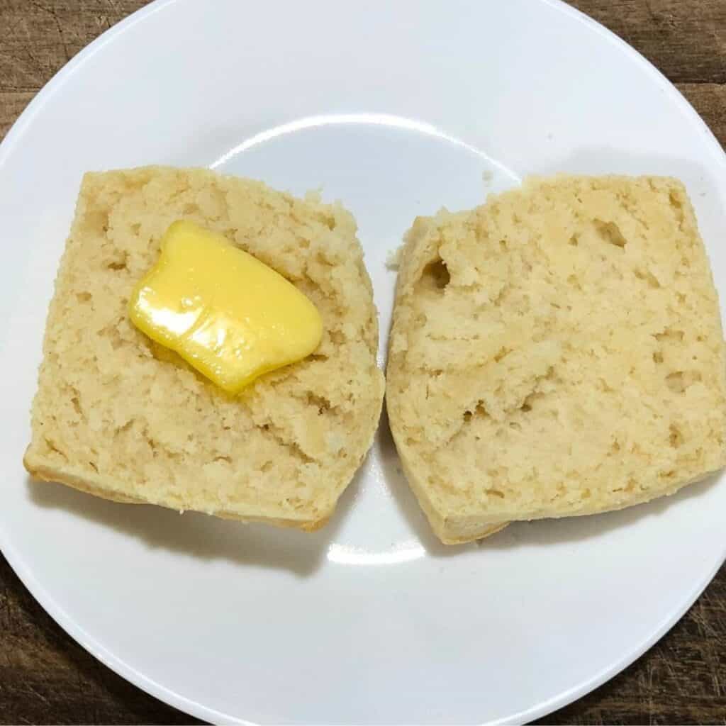 A homemade biscuit cut open on a white plate. The biscuit has a pat of butter melting on one of the halves.