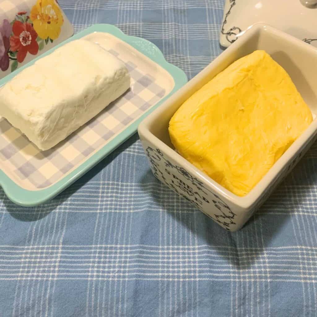 Homemade cream cheese on a ceramic blue and white checked butter dish next to homemade butter in a white ceramic butter dish. Both are on a blue and white checked kitchen towel.