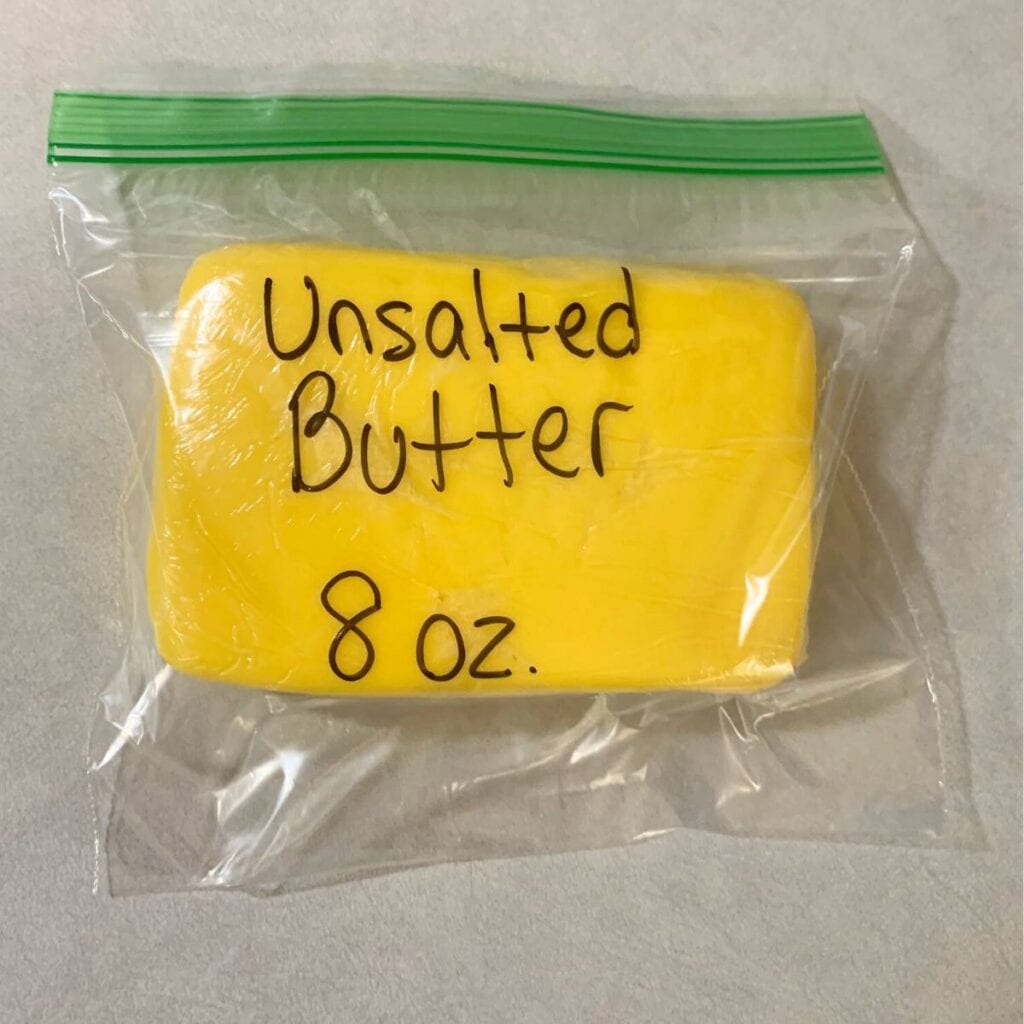 A block of yellow, homemade butter wrapped in plastic wrap and put inside a clear freezer bag. The freezer bag is labeled "unsalted butter" and eight ounces.