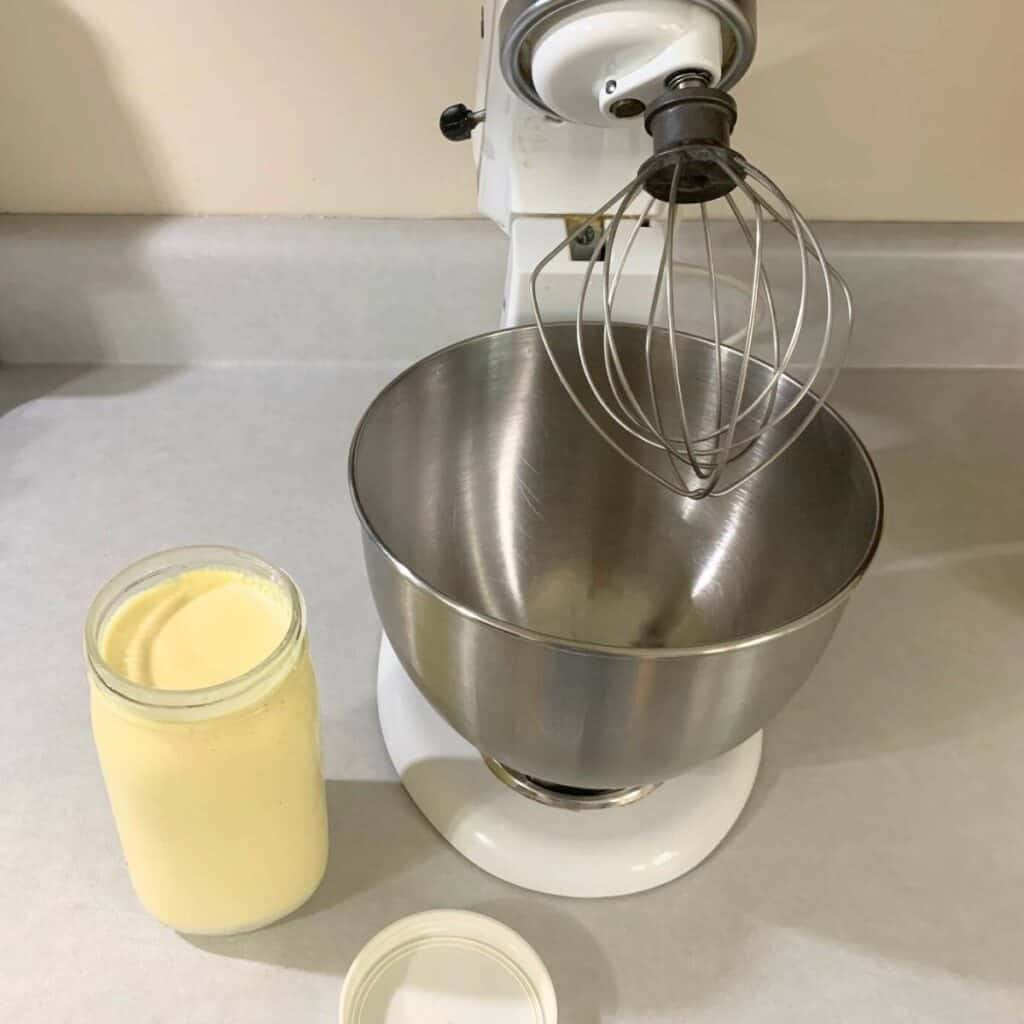 A stand mixer with stainless steel bowl and whisk attachment sitting on a kitchen counter. Next to it is a quart of heavy cream in a glass mason jar.