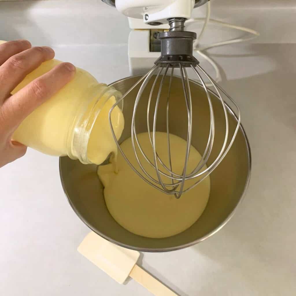 Woman pouring raw cream from a quart glass mason jar into the bowl of a white stand mixer with whisk attachment.