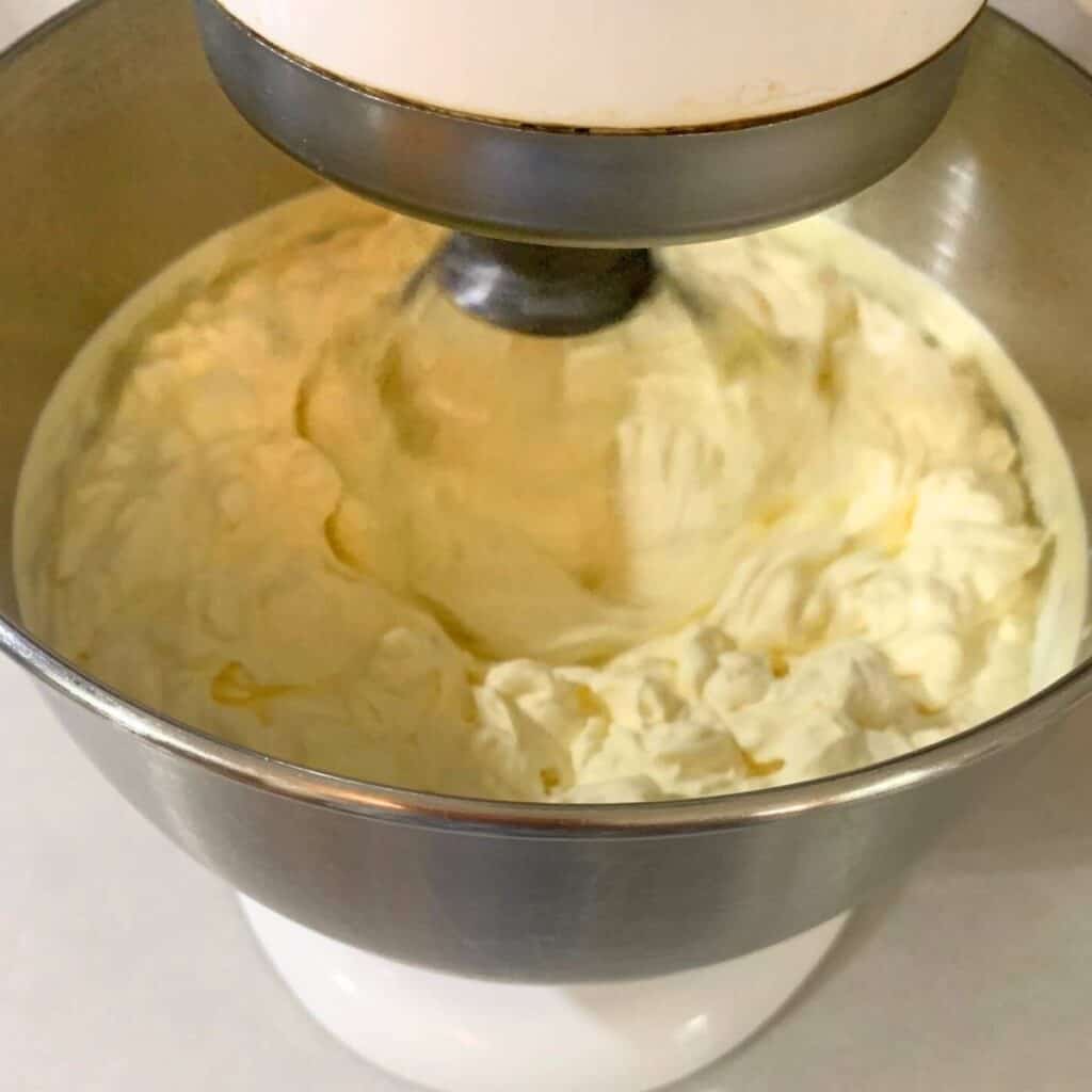 Raw cream in a white stand mixer with whisk attachment that is being whisked and has reached the "stiff peak" consistency.