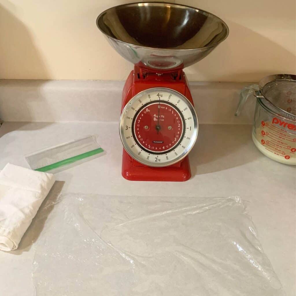 A red, mechanical kitchen scale, piece of plastic wrap, white flour sack towel, and freezer bag on a kitchen counter.