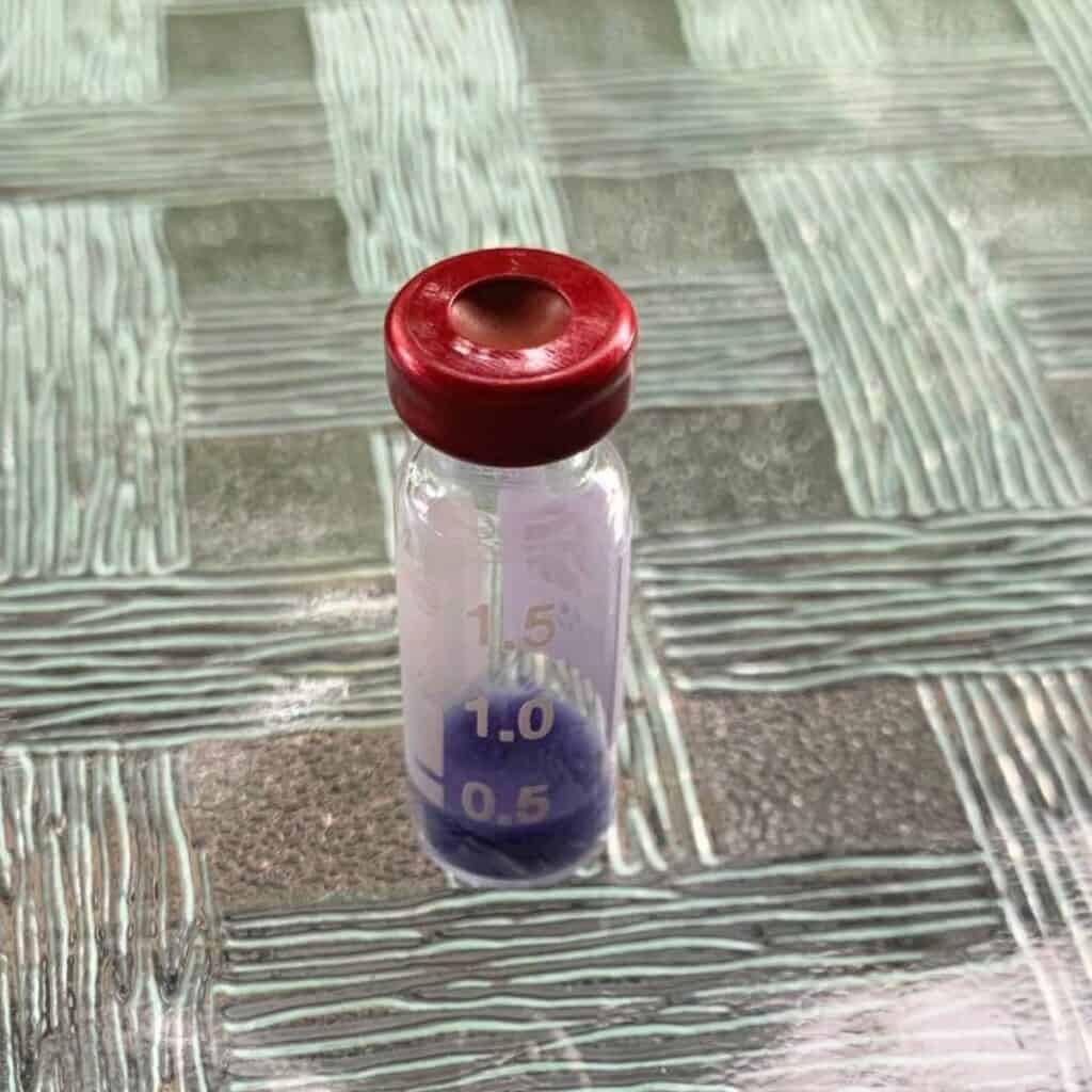 A glass vial showing dry blue powder at the bottom and a red lid. It is sitting outdoors on a glass table.