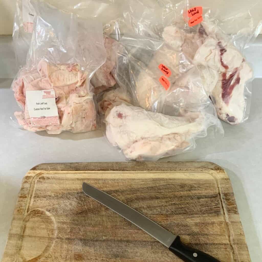 Packages of various pork fat cuttings from a butcher sitting on top of a kitchen counter next to a wooden cutting board and knife.