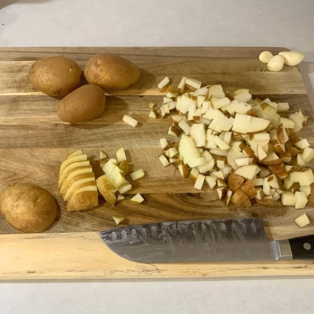 Washed russet potatoes where some have been diced small on a wooden cutting board next to a black chef knife.