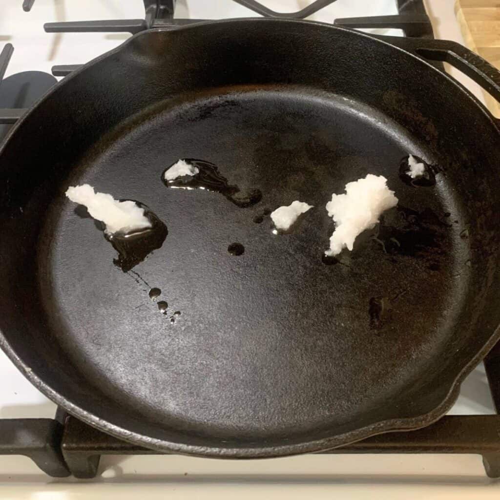 A few tablespoons of coconut oil in a cast iron skillet on the stove. The coconut oil is in the process of melting.