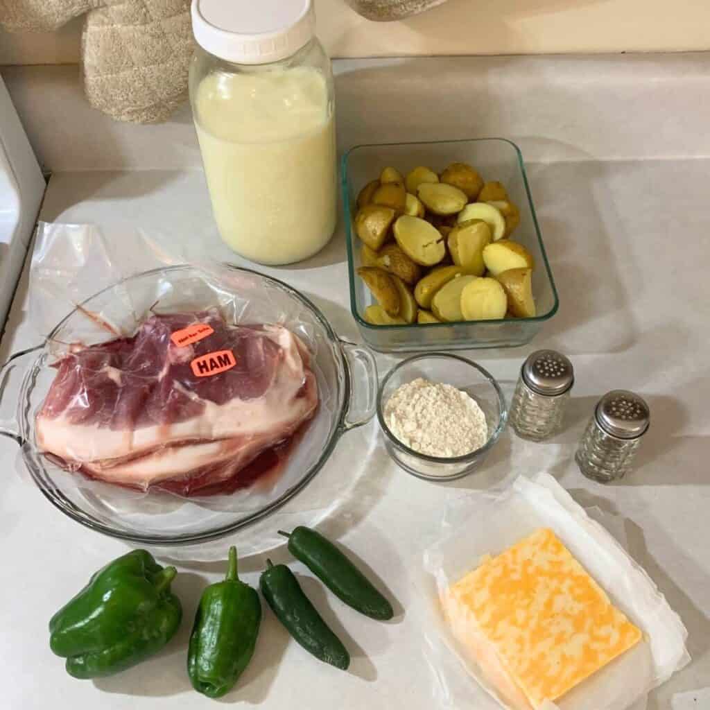 Ingredients for making ham, potato, and cheese casserole sitting on a kitchen counter. These include four green peppers, a ham steak thawing in a glass bowl, a half gallon mason jar of milk, a glass bowl with flour, a glass dish with cooked potatoes, a salt and pepper shaker, and sliced cheese.