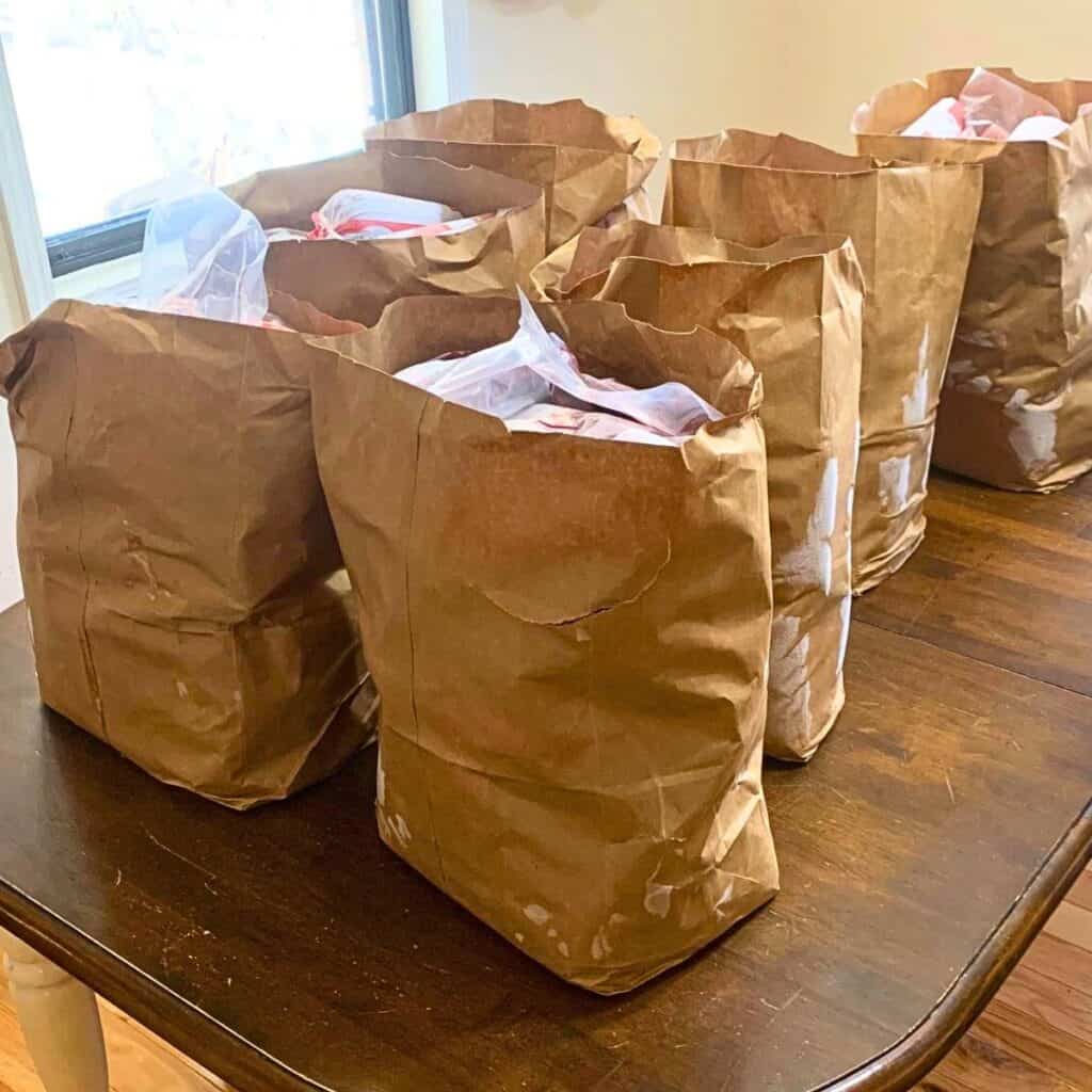 Seven brown paper grocery bags sitting on a wood kitchen table, and filled with frozen cuts of beef.