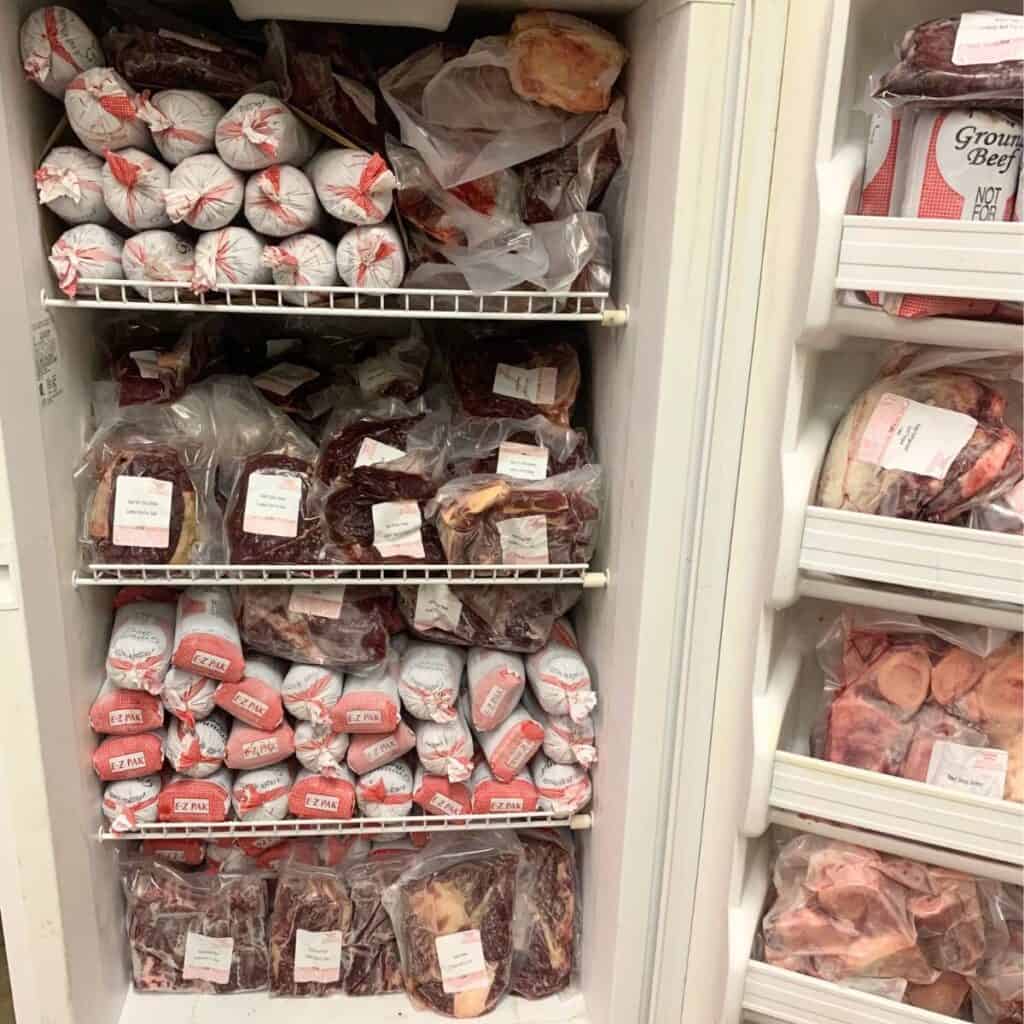 A white, upright freezer with the door open, showing the freezer is full of different cuts of frozen beef.