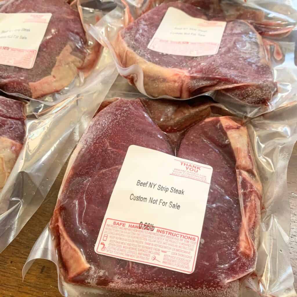 Several packages of frozen New York strip steaks on a wooden kitchen table.