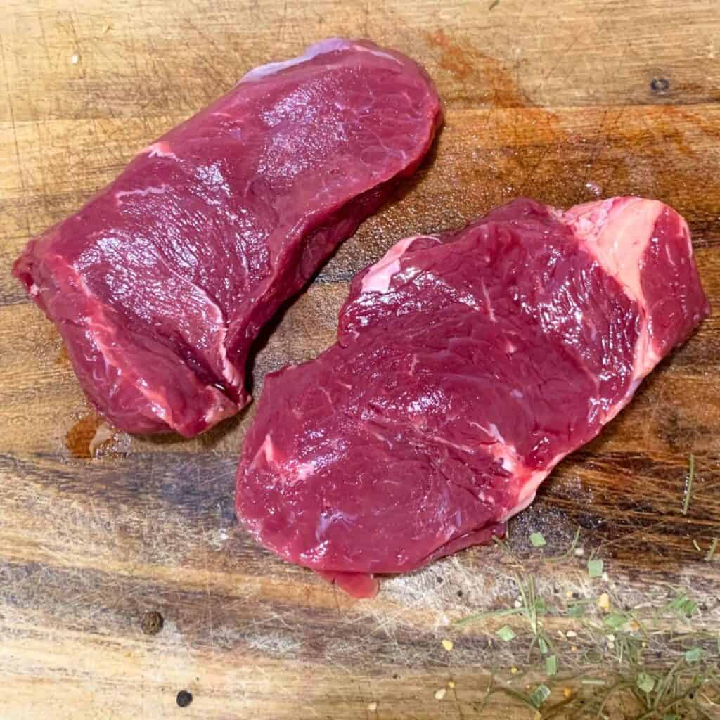 Two raw steaks on a wooden cutting board.