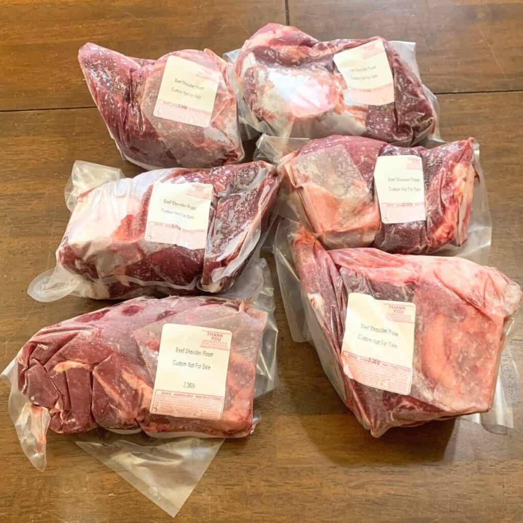 Six packages of frozen beef shoulder roast on a wooden kitchen table.